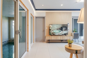 Una sala de estar moderna con un televisor de pantalla plana grande, un mueble de madera para el equipo audiovisual, una mesa redonda de madera con una planta y un ventanal que da a una zona de piscina.