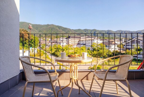 A balcony with two wicker chairs, a small round table, and a view of a town with buildings and hills in the background.