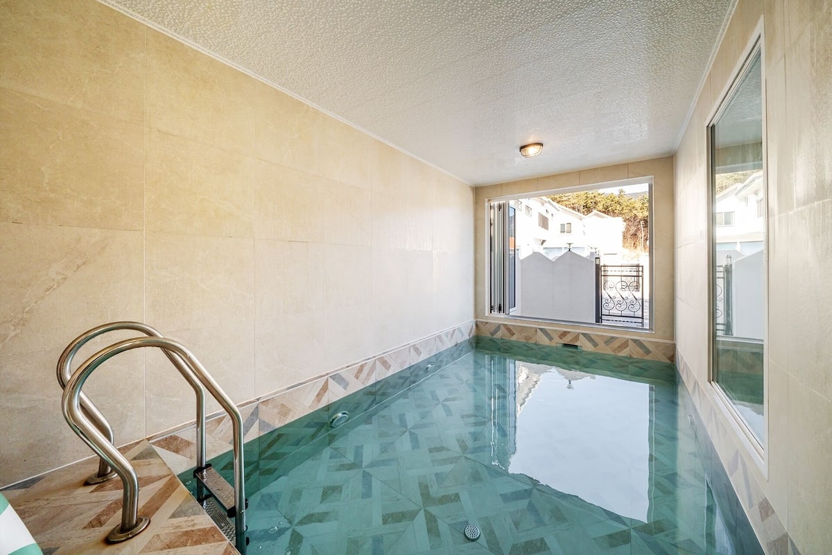 Une piscine dotée d’un plancher en verre, d’une rambarde métallique et offrant une vue sur un quartier résidentiel par le biais d’une fenêtre.