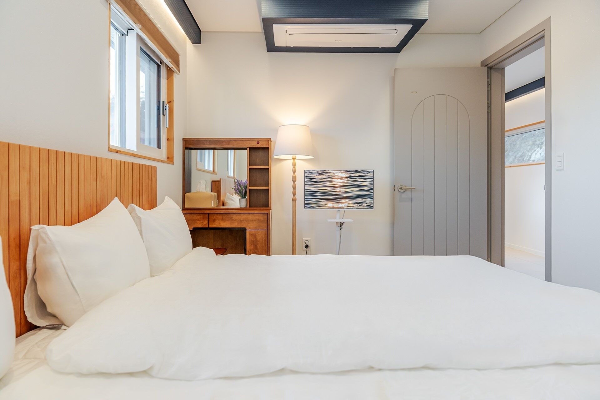 Un dormitorio con una cama grande, un cabecero de madera, una mesita de noche, un espejo, una lámpara de pared, una puerta y una ventana.