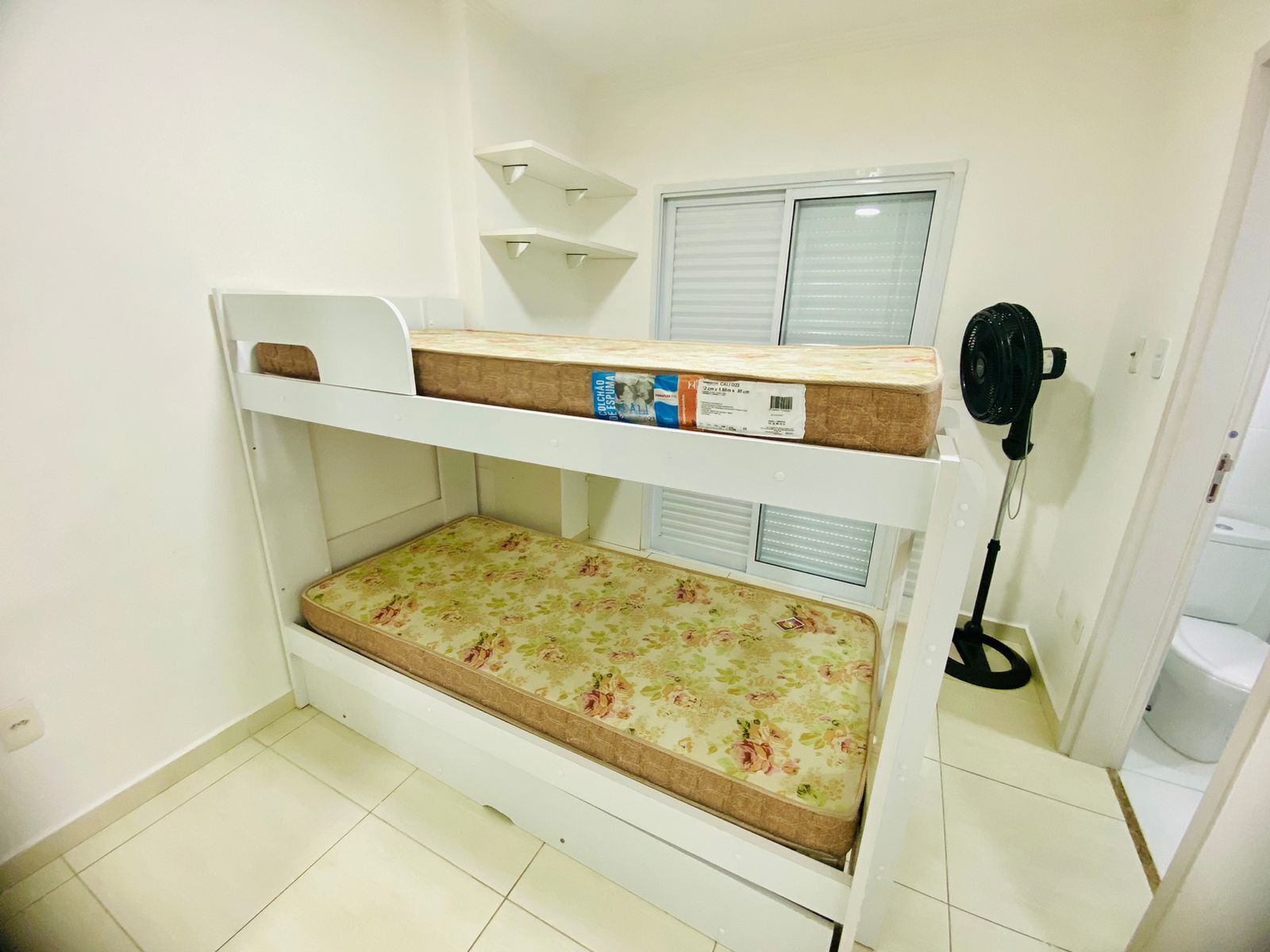 Ein Etagenbett mit Matratze und Kissen, ein kleiner Tisch, ein an der Wand befestigter Ventilator und ein Fenster mit Jalousien.