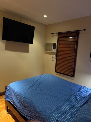 Une chambre à coucher comprenant un lit, un téléviseur fixé au mur, un appareil de climatisation et une fenêtre avec des stores.