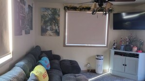 Ein Wohnzimmer mit Ecksofa, Deckenventilator, Flachbildfernseher und einem Fenster mit Jalousien.