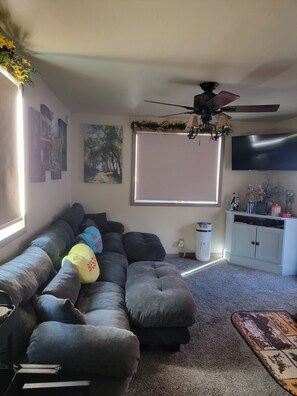 A living room with a sectional sofa, a ceiling fan, a flat-screen TV, and a window with blinds. - A Cozy Cabin You'll Want to Return (Big Bear)