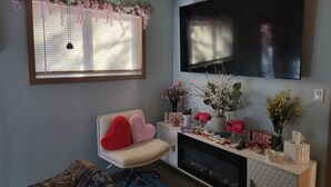 Ein Wohnzimmer mit an der Wand montiertem Fernseher, einer Couch, einem Sessel mit herzförmigen Kissen, einem Kamin und einem Fenster mit Jalousien.