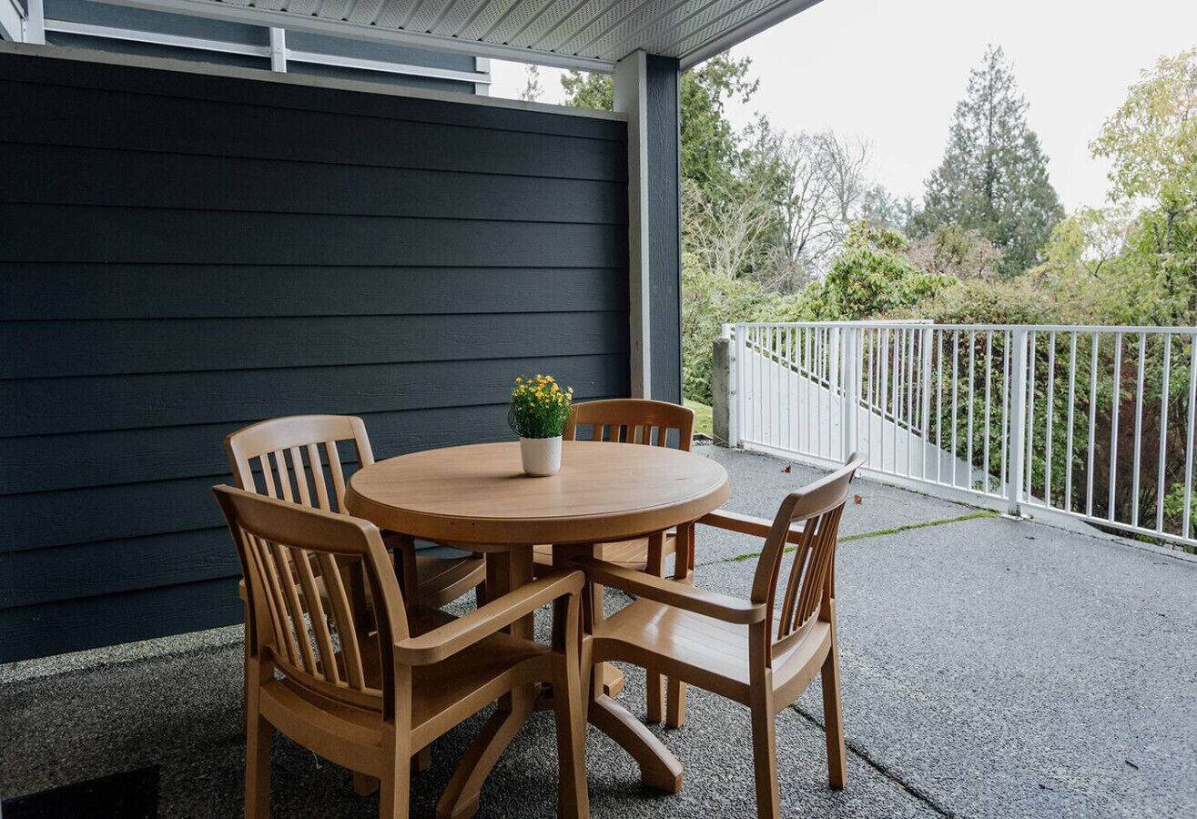 Eine überdachte Terrasse mit einem runden Holztisch und Stühlen, einem Blumentopf und einem weißen Geländer.