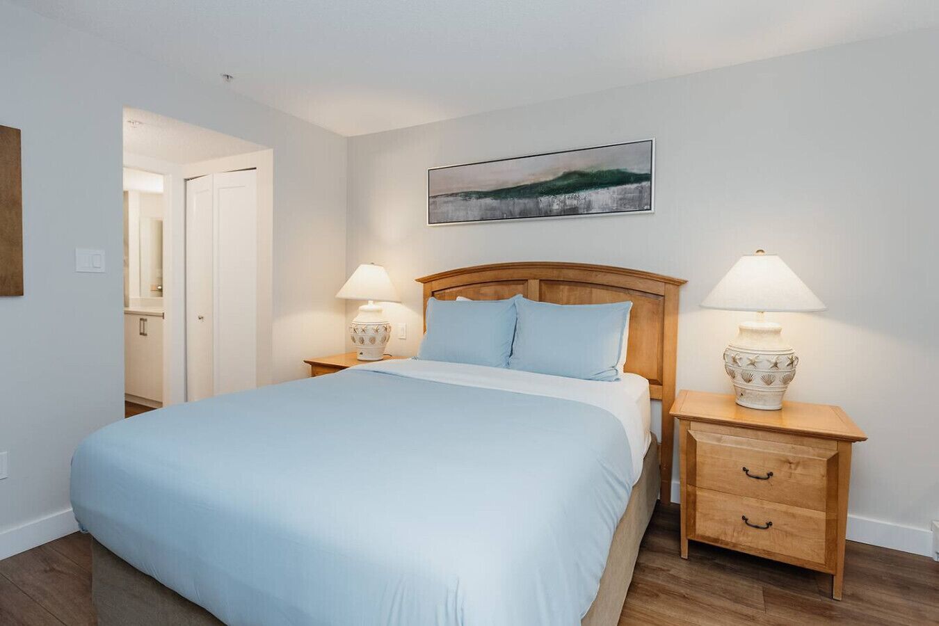 Une chambre à coucher avec un lit, deux tables de chevet avec des lampes, une tête de lit en bois, un tableau au mur et une porte donnant sur une salle de bain.