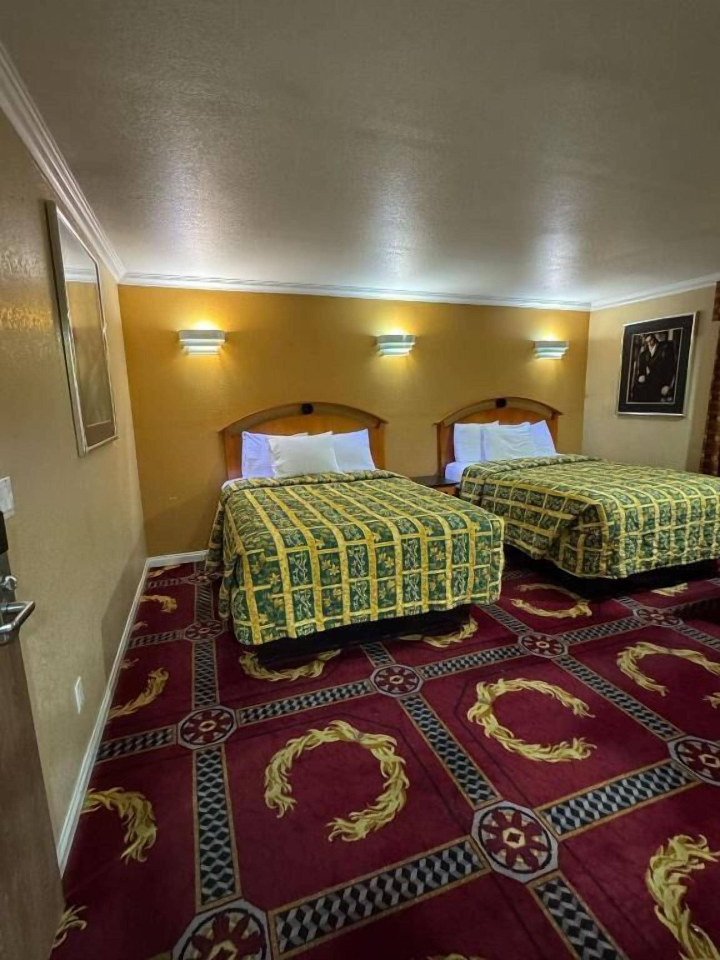 A hotel room with two beds, each with a patterned bedspread, a red carpet with geometric designs, and wall-mounted lights.