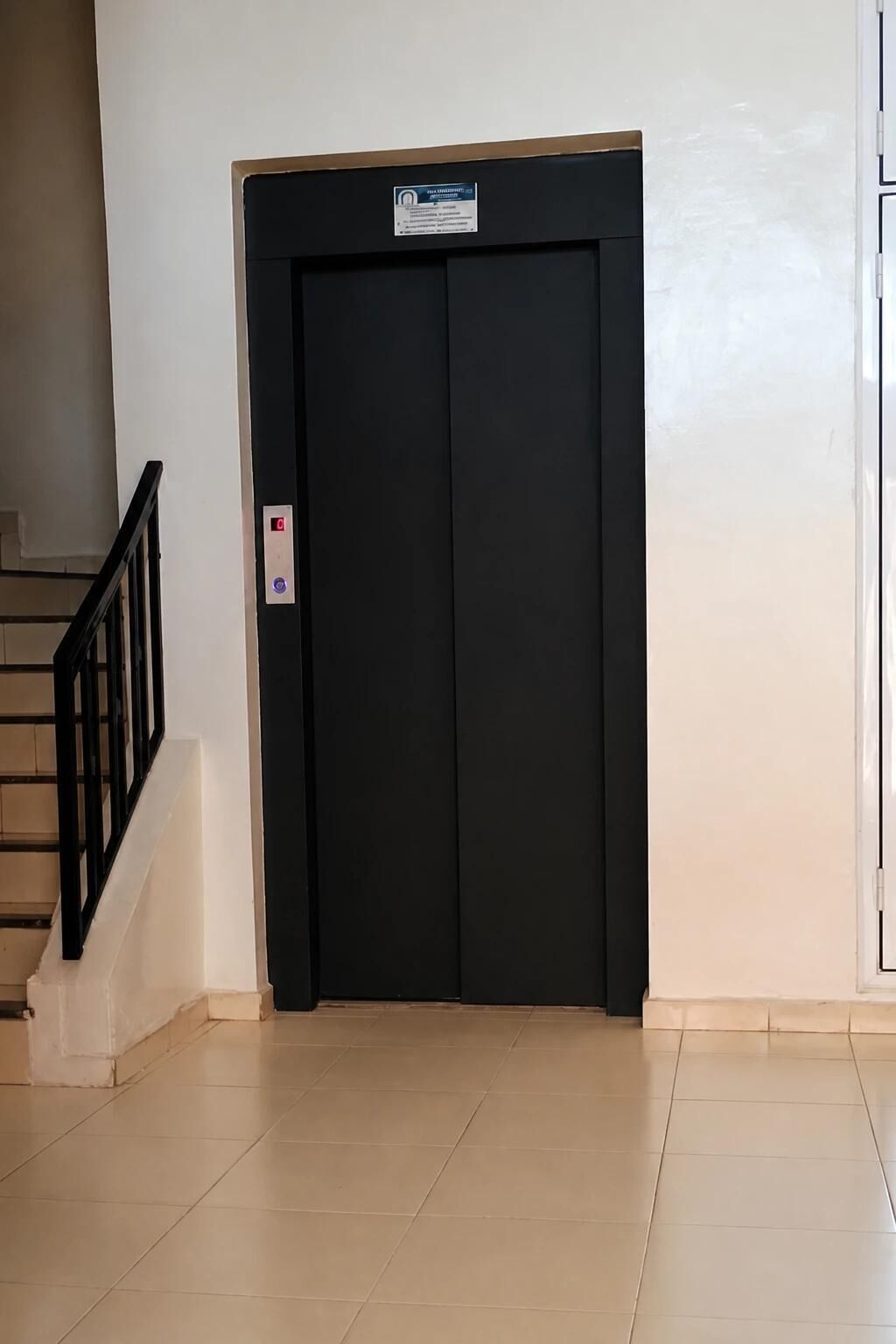 Una puerta de ascensor negra con un letrero encima, una escalera con barandilla negra a la izquierda y una pared lisa.