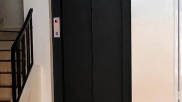 Una puerta de ascensor negra con un letrero encima, una escalera con barandilla negra a la izquierda y una pared lisa.