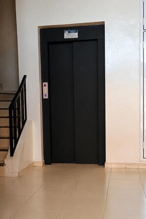 Een zwarte liftdeur met een bord erboven, een trap met een zwarre leuning links en een effen muur.
