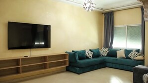 A living room with a large flat-screen TV mounted on the wall, a teal sectional sofa with patterned cushions, a wooden entertainment unit, and a decorative chandelier. - Modern stay with AC, Wi-Fi & parking near railway station (Fes)