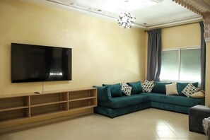 Una sala de estar con un televisor de pantalla plana grande montado en la pared, un sofá modular color turquesa con cojines estampados, un mueble de entretenimiento de madera y una lámpara de araña decorativa.