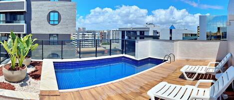 A rooftop pool area with a blue-tiled pool, wooden deck, and white lounge chairs.