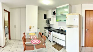 A compact kitchen with white cabinets, a microwave, a stove, and a refrigerator. There is a dining table with chairs and a small rug. - Vacanze - Port Ville II (Maceió)