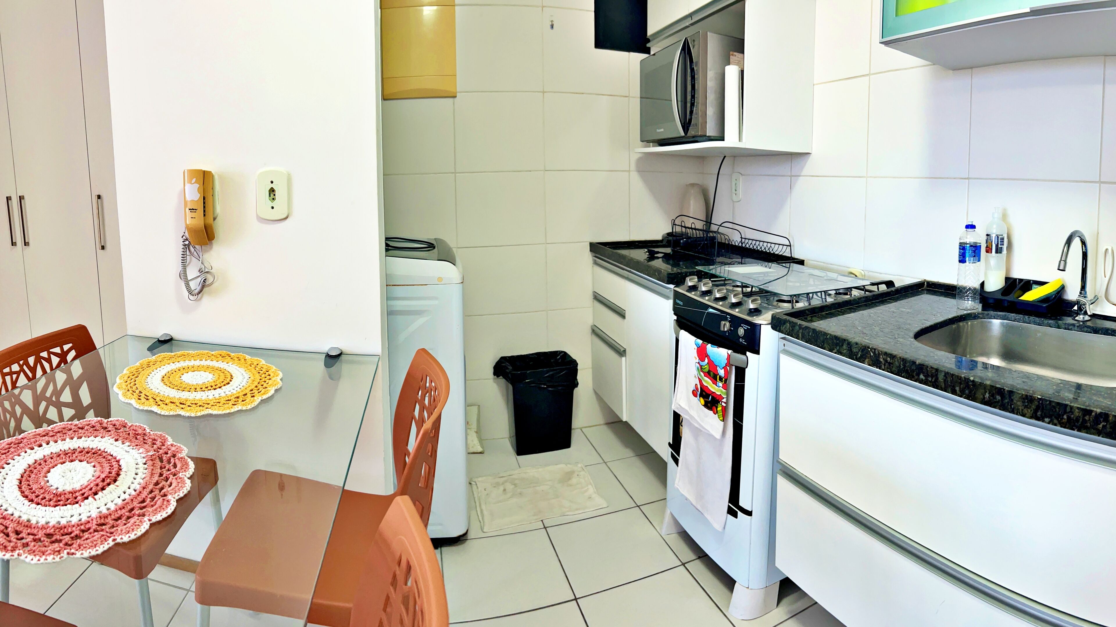 Una cocina moderna con armarios blancos, un lavamanos de acero inoxidable y una estufa a gas. Hay una mesa de comedor con un mantel individual de crochet y dos sillas.
