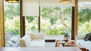 Un espace de vie moderne, à aire ouverte, doté d’une grande fenêtre offrant une vue sur une végétation luxuriante. On y trouve deux canapés blancs, une table basse en bois et un luminaire suspendu.
