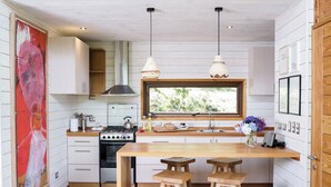 Une cuisine compacte avec des meubles blancs, un plan de travail en bois, une cuisinière à gaz et une fenêtre donnant sur un espace verdoyant. On y trouve deux tabourets en bois et une table.