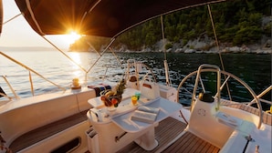Un velero con zona de comedor, que incluye una mesa con bebidas y un libro, frente a un atardecer y una costa rocosa.