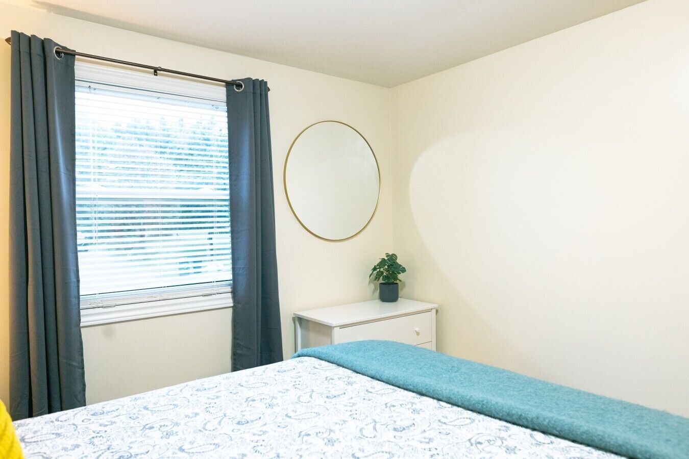 A bedroom with a bed, a bedside table with a plant, a window with curtains, and a wall-mounted mirror.