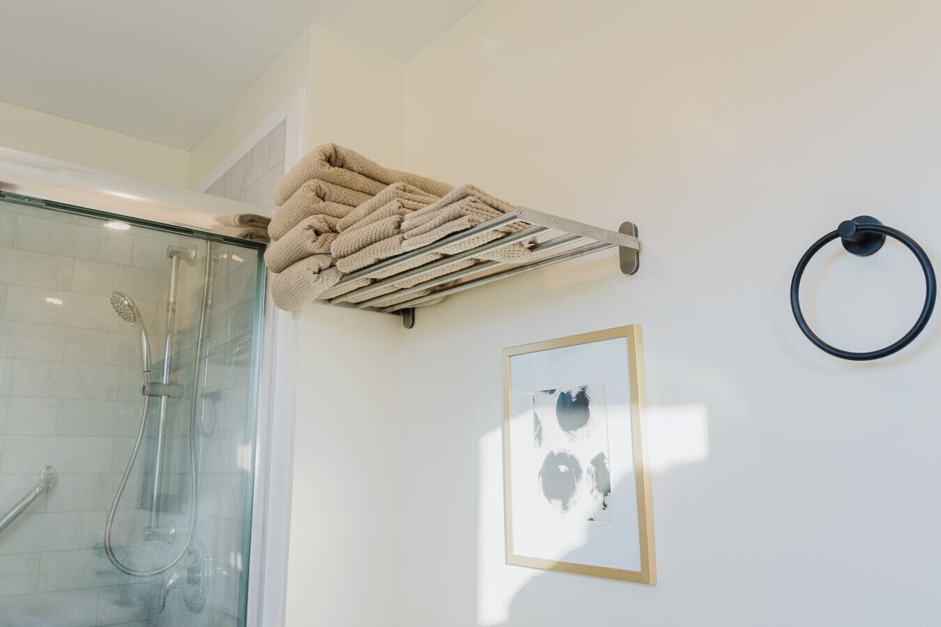 A bathroom with a glass shower enclosure, a towel rack with folded towels, a framed abstract artwork, and a black circular object on the wall.