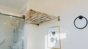 A bathroom with a glass shower enclosure, a towel rack with folded towels, a framed abstract artwork, and a black circular object on the wall.