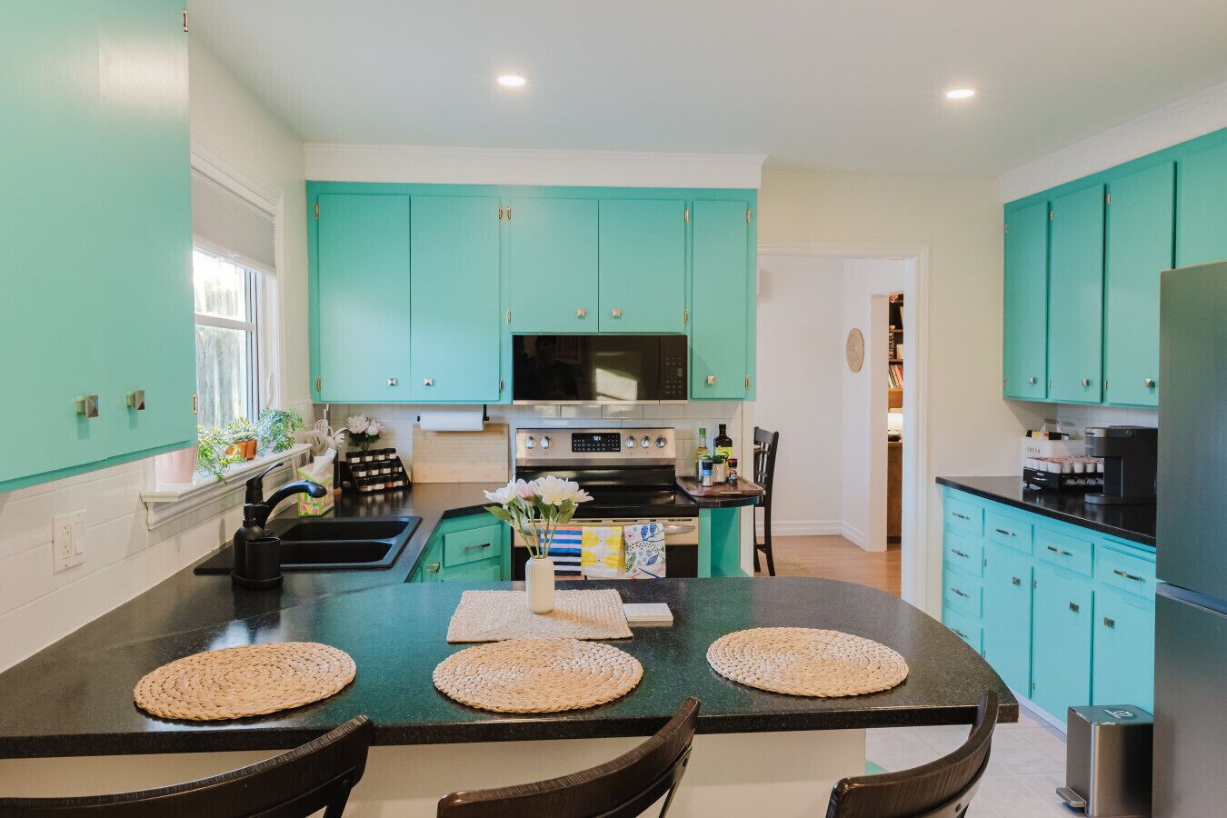 A kitchen with teal cabinets, a central island with woven placemats, and a dining area with chairs.
