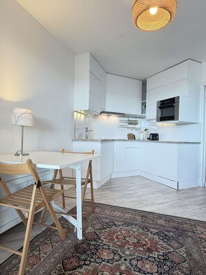 A modern kitchen with white cabinetry, a dining table with chairs, a patterned rug, and a hanging light fixture. - PORNICHET - STUDIO CABINE VUE MER (PORNICHET)