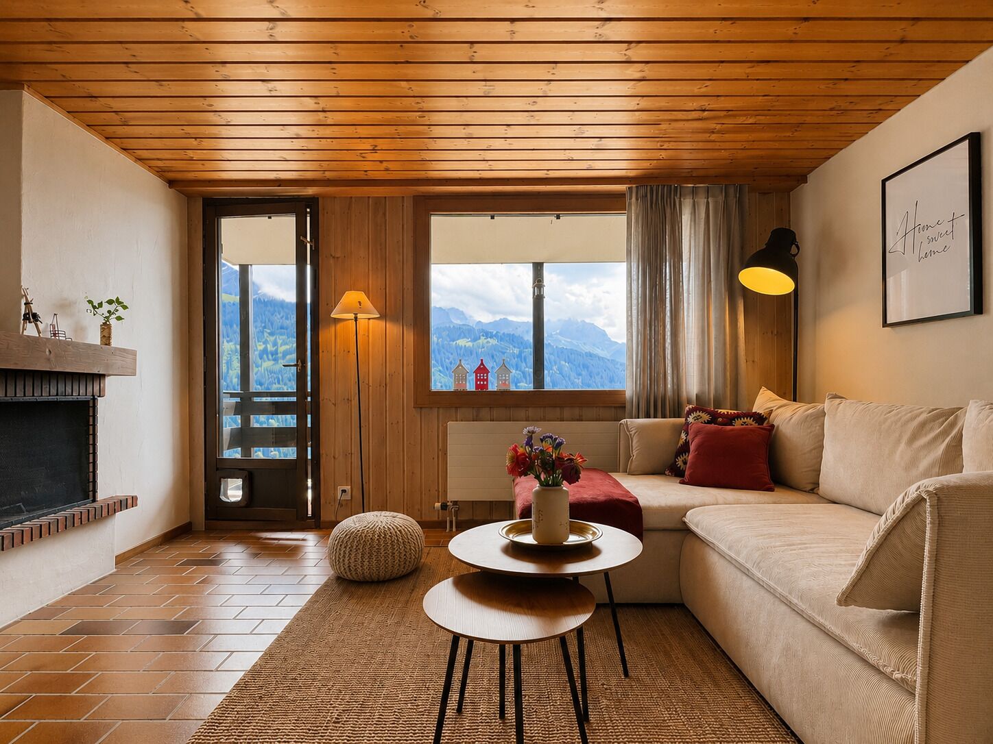 Un salon chaleureux avec une cheminée, un canapé, une table ronde ornée d’un vase de fleurs et une vue sur les montagnes par la fenêtre.