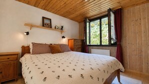 Une chambre avec un plafond en bois, un lit recouvert d’une literie à motifs floraux, une table de chevet, une commode, une fenêtre avec des rideaux et une étagère sur laquelle se trouvent un tableau et une lampe.