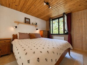 Ein Schlafzimmer mit Holzboden, einem Bett mit Blumenmuster, einem Nachttisch, einer Kommode, einem Fenster mit Vorhängen und einem Regal mit einem Bild und einer Lampe.