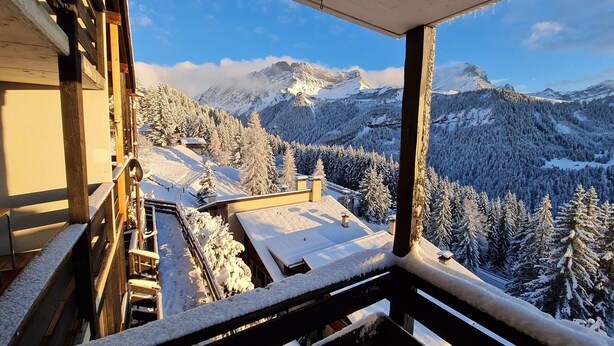 Un paysage de montagne enneigé, avec un balcon en bois et une vue sur des arbres et des montagnes couverts de neige.