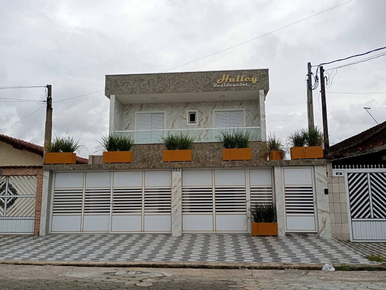 Un edificio residencial con una placa que dice 'Halley Residential'. Cuenta con un acceso empedrado con un diseño a cuadros, varias puertas de garaje y plantas en macetas en la fachada.
