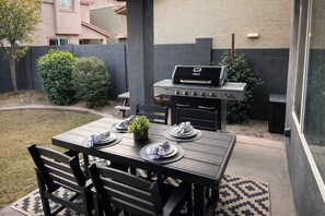 Eine Außenpatio mit einem Holztisch für zwei Personen, einem Grill und einem kleinen Gartenbereich.