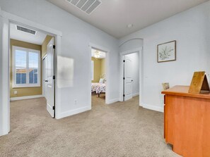Ein geräumiger Raum mit Teppichböden, weißen Wänden und einem Holztisch. Mehrere Türen führen in andere Räume, und ein Fenster mit Jalousien ist vorhanden.