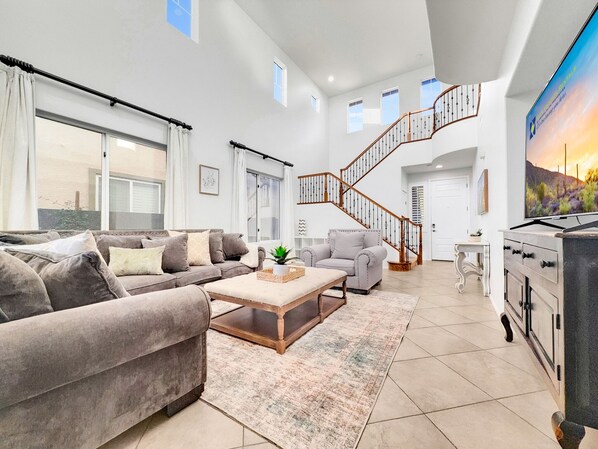 Ein geräumiges Wohnzimmer mit einem grauen Sofa, einem kleineren Sessel, einem Couchtisch und einer Treppe, die eine weitere Ebene hinaufführt.