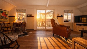 A spacious living room with a large sliding glass door leading to a balcony, wooden flooring, a leather armchair, a dining area with chairs, and a television on a wooden cabinet. - Clam Lake Waterfront Cottage w/ Kayaks (Bellaire)