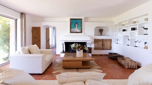 Amplia sala de estar con chimenea, sofás blancos, una mesa de centro con flores y vista a zonas verdes.