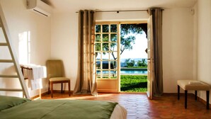 Un dormitorio con cama, una silla, una mesita y vistas a una piscina y follaje a través de un ventanal amplio.
