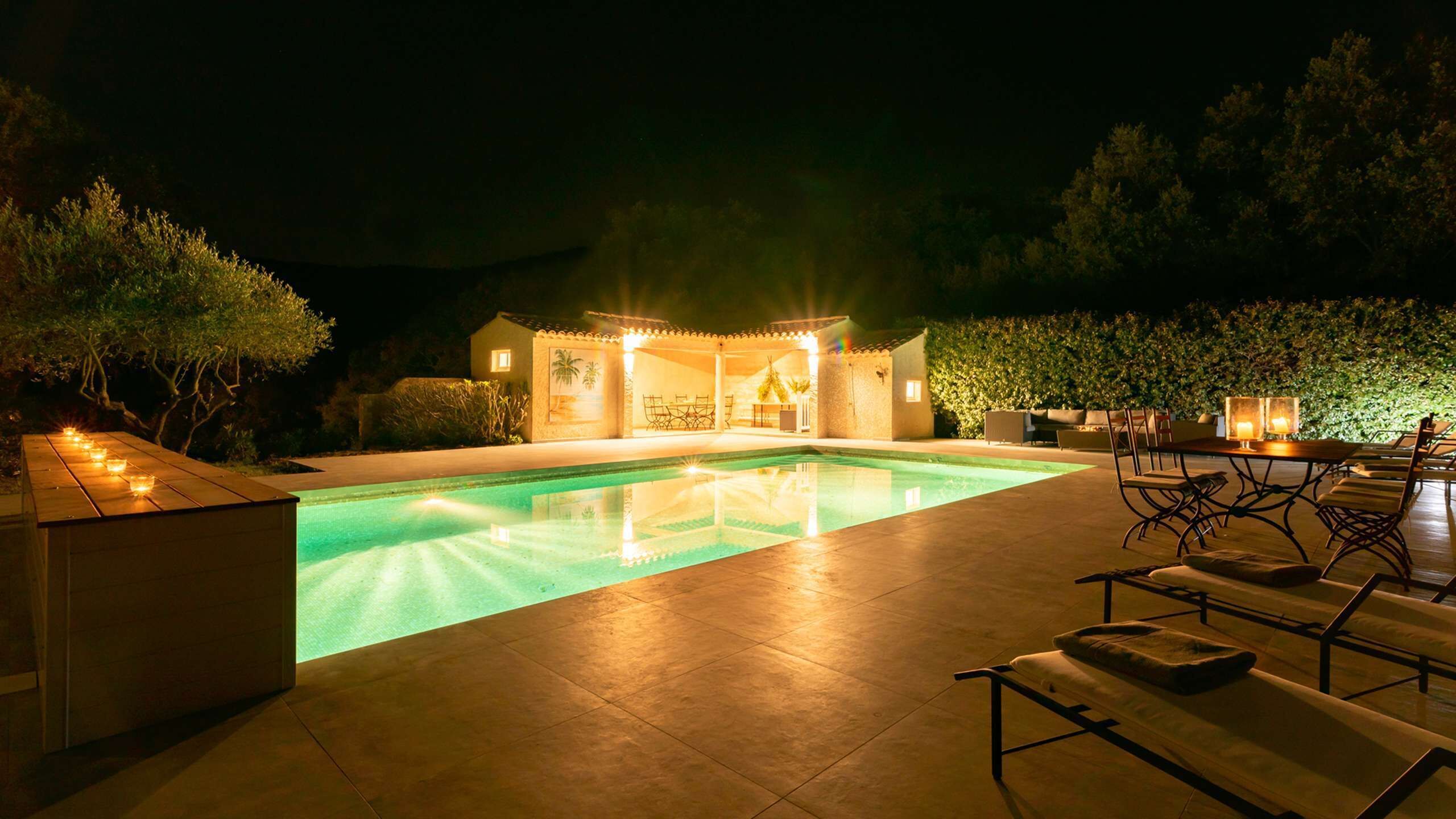 A nighttime outdoor pool area with a covered seating area, a table, and a small building with a mural.