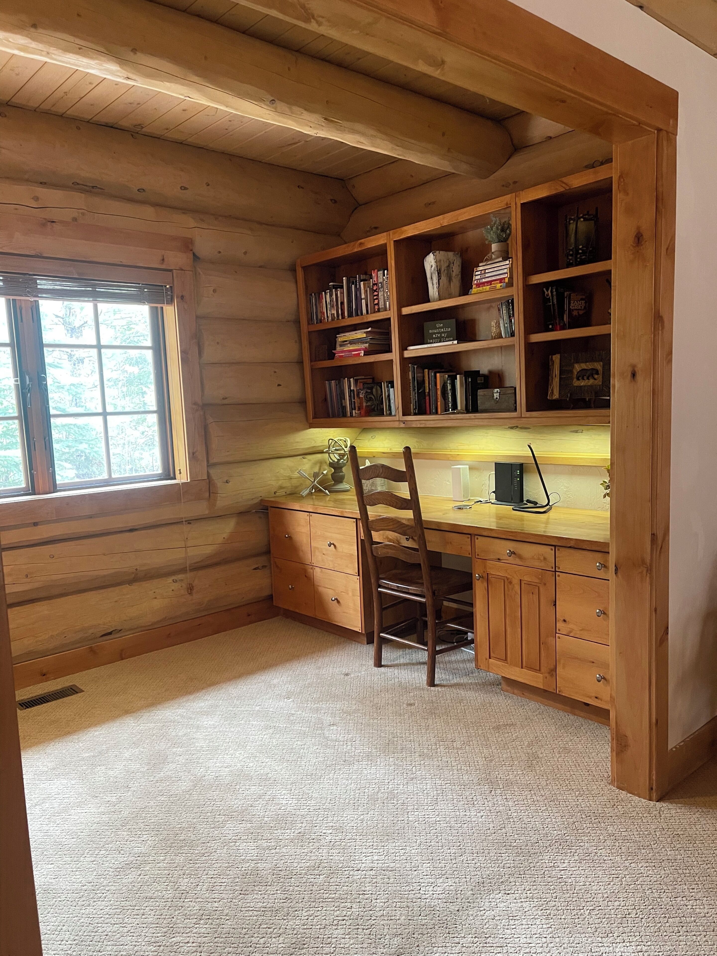 Interior de una cabaña de madera con un escritorio, estanterías y una ventana.