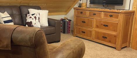 A loft-style room with a brown leather sofa, a wooden dresser, and a flat-screen TV mounted on the wall.