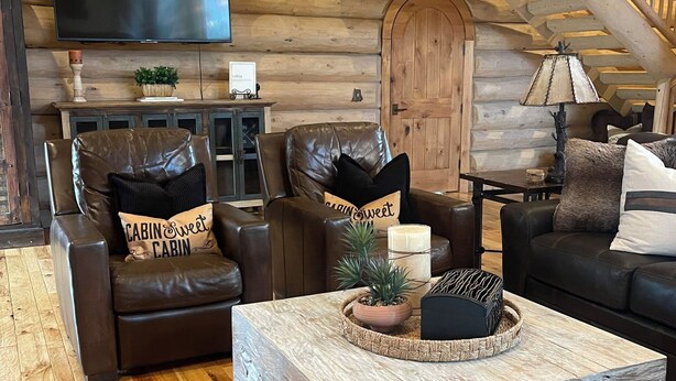 A log cabin interior with a wooden staircase, leather sofas, a flat-screen TV, and a rustic coffee table.