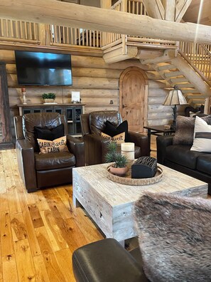 A log cabin interior with a wooden staircase, leather sofas, a flat-screen TV, and a rustic coffee table. - Moose Hollow Rustic Log Cabin Rustic Weber River Retreat in Oakley, UT Sleeps 22 (Oakley)