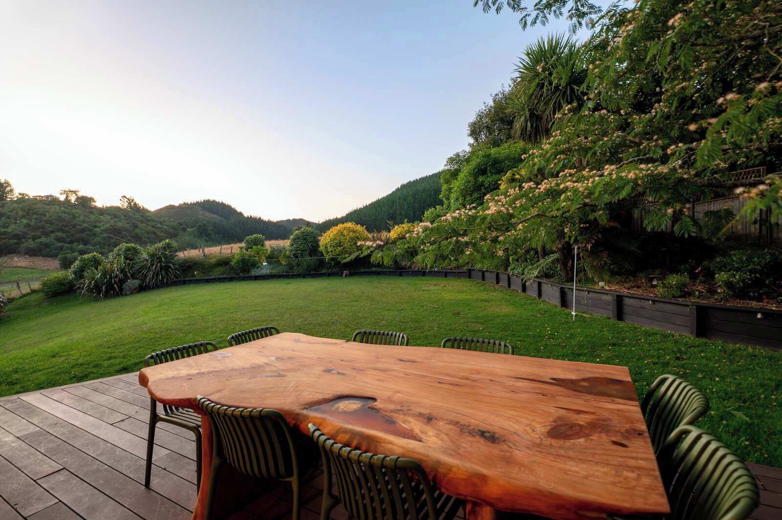 A wooden deck with a table and chairs, surrounded by a lush garden and a view of a hilly landscape.