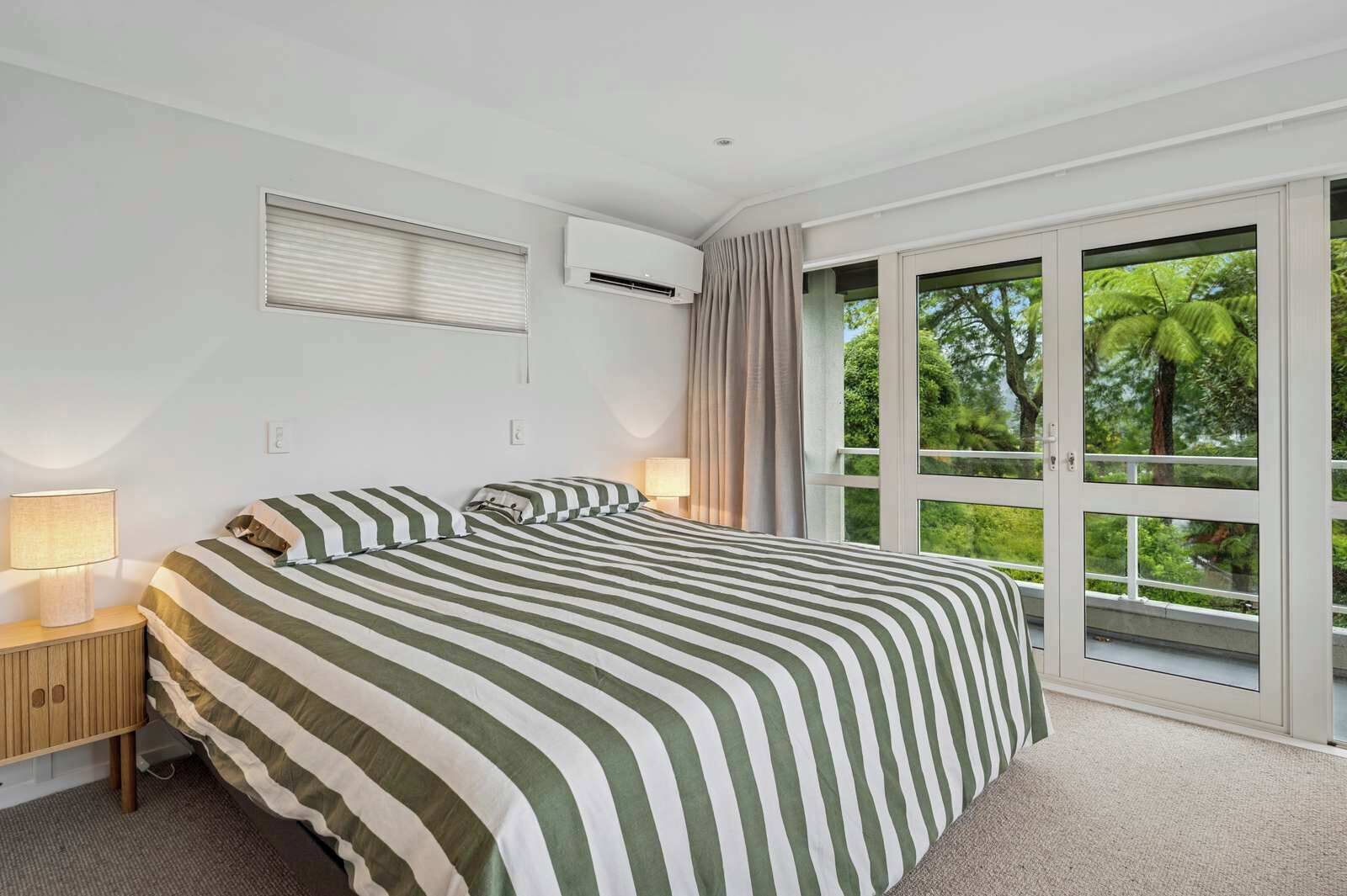 A bedroom with a large bed, a nightstand, an air conditioning unit, and a sliding glass door to a balcony with a view of trees.