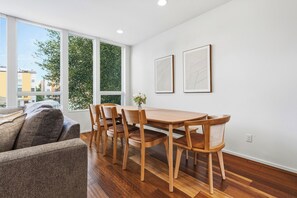 Un salon moderne avec une table à manger en bois, quatre chaises, un canapé et de grandes fenêtres donnant sur des immeubles et des arbres.