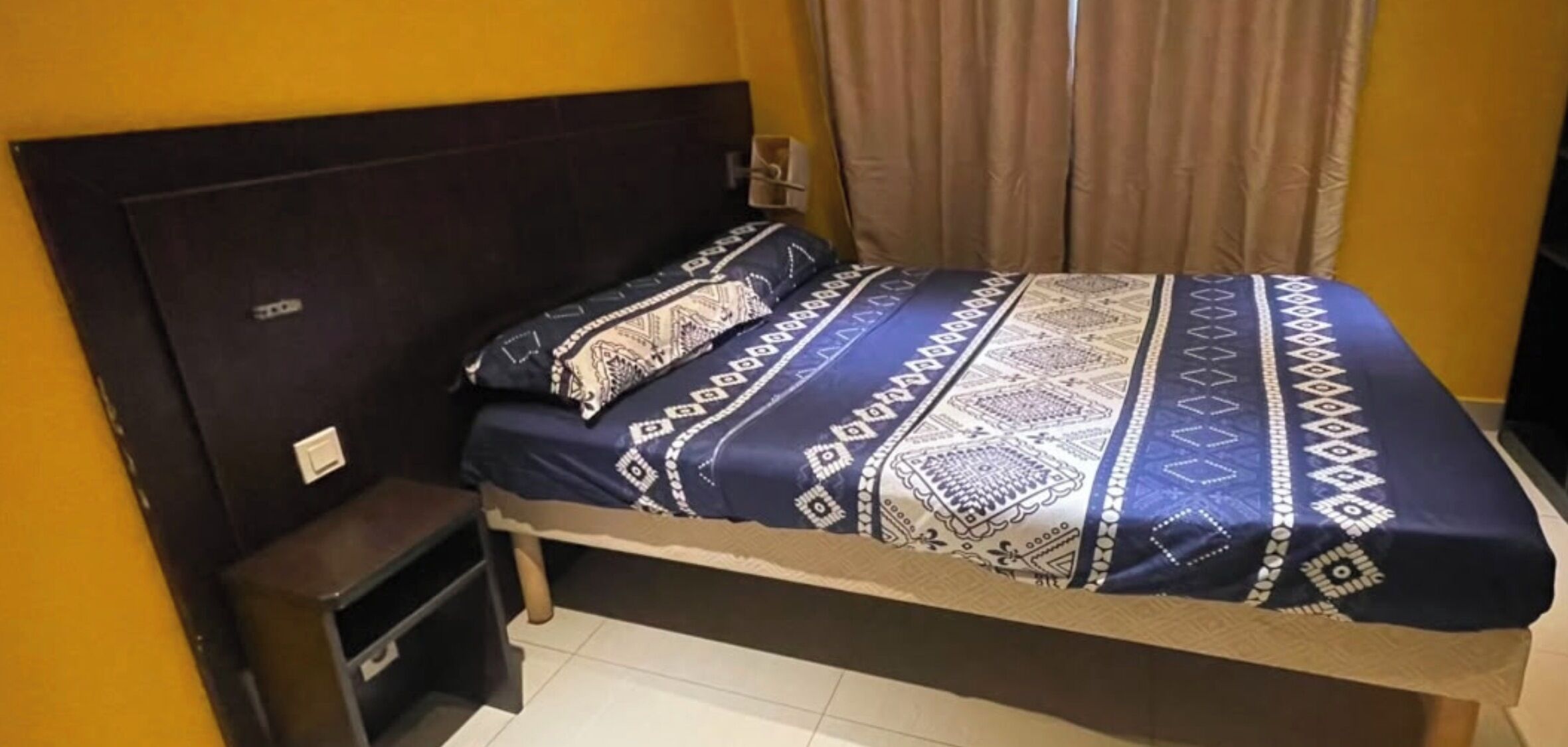 A single bed with a patterned bedspread and pillows, a nightstand, and a wall-mounted light switch.
