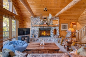 A log cabin interior with a stone fireplace, wooden beams, a television, and a cozy seating area.