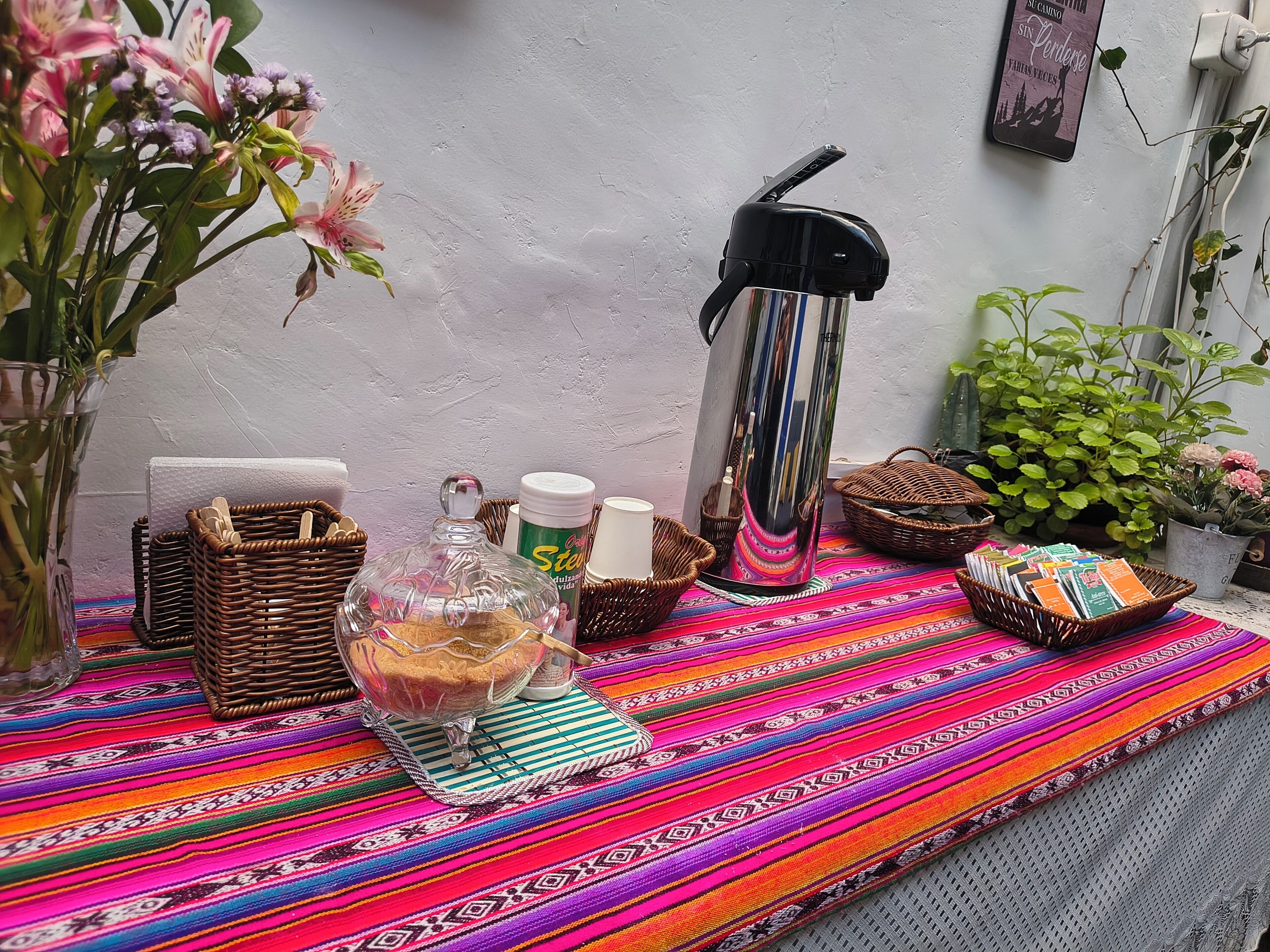 A table with a colorful woven tablecloth, a glass decanter, a thermos, and a basket of books.